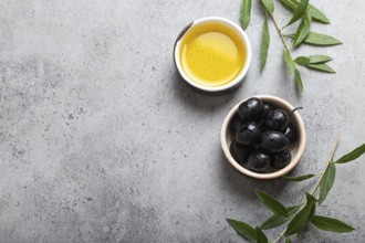 Black olives and extra virgin olive oil in little bowl with olive tree branches top view on gray
