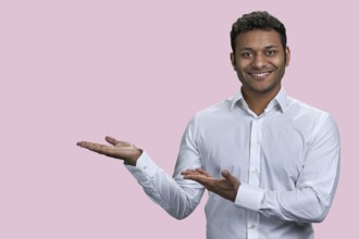 Happy positive young indian man is showing copy space with both hands. Isolated on pink background