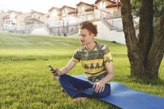 Young stylish handsome guy is sitting on grass and talking on the phone resting in the park. Taking