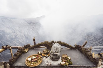 Traditional offerings and a statue on an altar with a misty volcano background, Bromo volcano, Sri