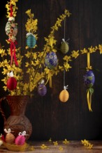 Branches of forsythia (Forsythia) in a rustic jug hung with Easter eggs against a dark background