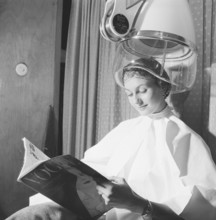 Hair dryer in the 1950s. A woman is sitting comfortably and reading Vogue magazine, while drying