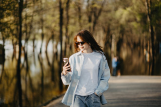 A woman wearing sunglasses, looking at her phone while walking in a sunlit park