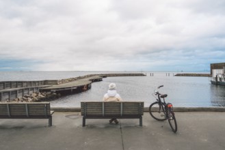 A young Caucasian woman sits with her back on a wooden bench overlooking the Baltic Sea on the