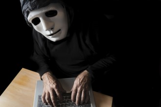 Hacker dressed in black with mask and hood typing on a computer while looking at the camera