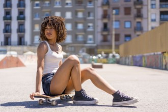 Stylish young woman taking a break, sitting on her skateboard in a skate park, enjoying the urban