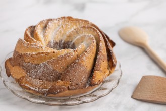 Fresh marble bundt cake, cake with icing sugar next to a rustic wooden spoon on a light-coloured