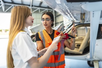 Female aircraft maintenance engineer wearing high visibility vest explaining airplane part to pilot