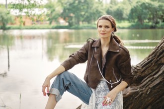 A woman wearing a brown jacket and light blue jeans sits on a tree trunk by a peaceful lake. The