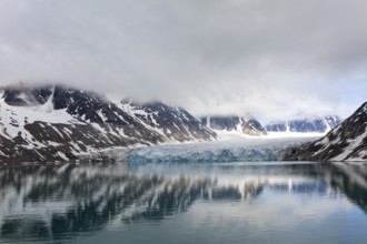 An impressive lake reflects the surrounding glaciers and mountains, surrounded by clouds and a calm
