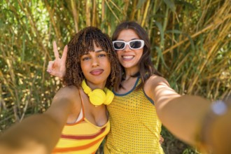 Two happy young women with headphones and sunglasses taking a selfie in a bamboo forest, one of