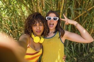 Two girls with headphones and sunglasses taking a selfie in a bamboo forest, making victory signs