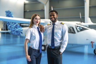 Two smiling airline pilots posing confidently in front of a private jet inside a spacious hangar,