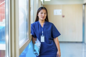 Confident young medic holding a clipboard and wearing blue scrubs is standing next to a window in a