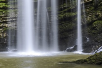 A waterfall with a stream of water flowing out of it. The water is brown and the rocks are green.