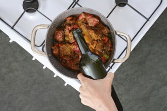 A hand adds a bottle of wine to a pot filled with meat and vegetables simmering on the stove. The