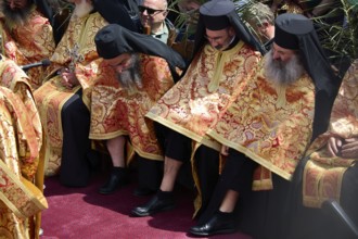 Orthodox priests in elaborate golden robes sit in the middle of a crowd during a ceremony, Orthodox