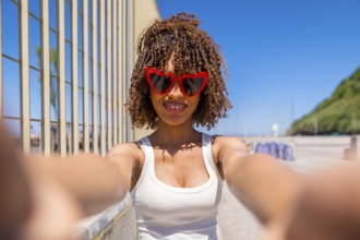 Smiling young woman with curly hair taking a selfie with heart shaped sunglasses on a sunny beach