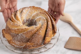 Hands present a bundt cake dusted with icing sugar on a glass plate