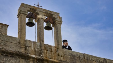 Priest next to bells on a wall in front of a blue sky, Monastery of St John the Theologian,