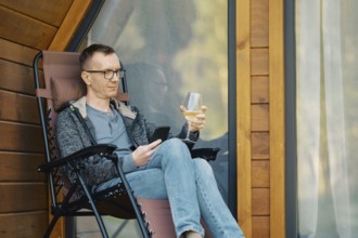 Middle-aged man looking at smartphone sitting on terrace of a house with a glass of wine