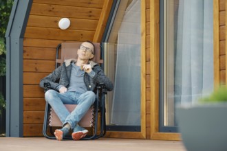 Middle-aged man enjoys a cigar while lounging in a chair on the terrace of a log cabin