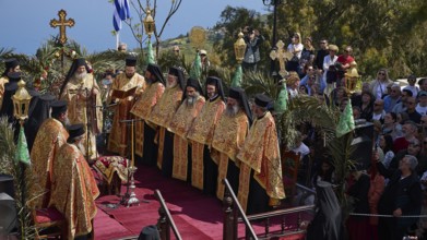 Priests in golden robes lead a religious ceremony on a decorated stage in front of a Greek flag and