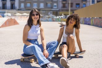 Two young women are sitting on their skateboards in a skate park, enjoying a break and smiling