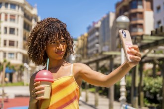 Young woman taking a selfie with her smartphone while holding a milkshake, enjoying a sunny day in