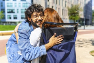 Young man with glasses is embracing a red haired woman sitting in a wheelchair on a sunny day in