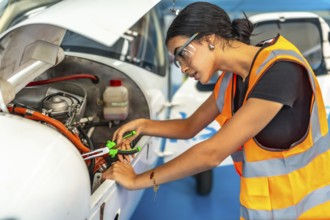 Young woman aircraft mechanic wearing safety glasses and high visibility vest using pliers while