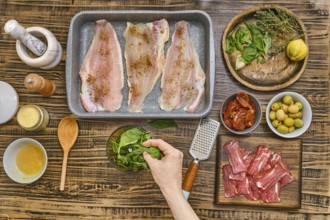 Fresh herbs, vegetables, and fish fillets are arranged on a wooden table. A hand adds spinach to a