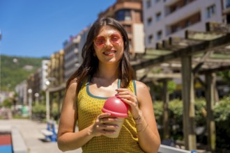 Happy young woman wearing sunglasses enjoying a refreshing drink on a sunny summer day in the city