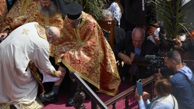 A priest in a golden robe blesses another, surrounded by cameras and a crowd, Orthodox ceremony,