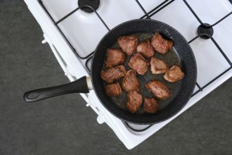 Diced beef is sizzling in a black skillet on a white stove. The meat is lightly browned and ready
