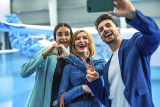 Three happy friends are taking a selfie in a hangar, making heart and victory signs, celebrating