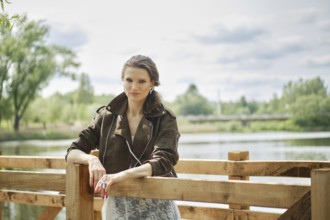 A woman stands confidently by a wooden railing in an elegant outfit, enjoying a peaceful afternoon