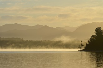 A calm lake at dawn with mist, surrounded by silhouetted mountains, Lake Mahinapua, Ruatapu, South