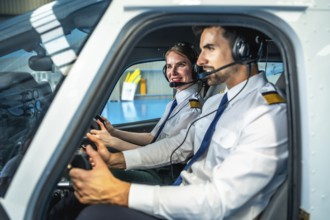 Male and female pilots, wearing headsets and smiling, operating a small airplane inside a hangar,