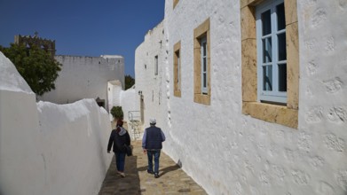 Two people walking along white buildings with windows on a sunny day, Mantomata Stones, Simandiri
