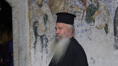 Priest in clerical garb in front of a religious mural, Monastery of St John the Theologian,