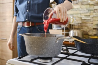 A person pours tomato sauce from a jar into a pot on the stove. The kitchen features modern decor