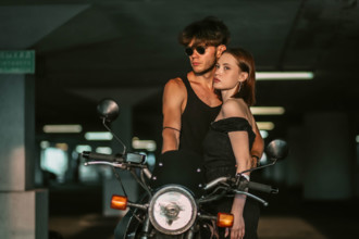 A couple in stylish outfits embraces near a motorcycle in a garage