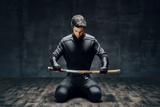 Warrior man sitting on floor posing with a sword over black background