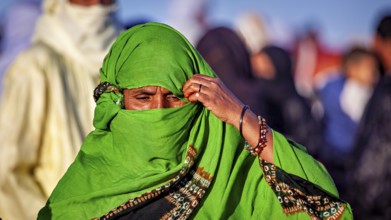 A woman in a green robe and jewellery pensively covers her face, Veiled woman at the Tamanrasett