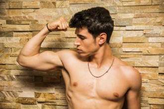 Attractive and muscular shirtless young man leaning against stone wall doing bicep pose