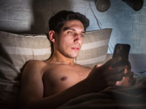 Handsome young man in bed typing on cell phone, sending text message or dialing number
