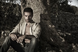 Ruggedly Handsome Man Seated at Base of Tree with Shirt Unbuttoned looking at camera, Relaxing in