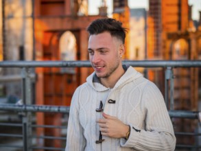Handsome trendy young man, smiling, standing on a sidewalk in city setting at sunset wearing a