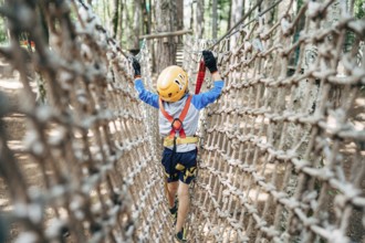 Boy walks across a mesh bridge, holding onto a cable. Back view.